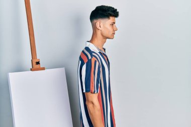 Young hispanic man standing close to empty canvas looking to side, relax profile pose with natural face with confident smile. 