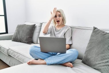 Young caucasian woman using laptop at home sitting on the sofa surprised with hand on head for mistake, remember error. forgot, bad memory concept. 