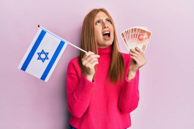 Young irish woman holding israel flag and shekels banknotes angry and mad screaming frustrated and furious, shouting with anger looking up. 