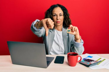 Beautiful middle age woman holding cpu computer processor with angry face, negative sign showing dislike with thumbs down, rejection concept 