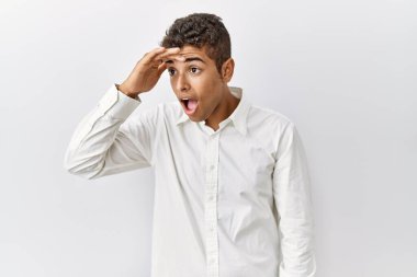 Young handsome hispanic man standing over isolated background very happy and smiling looking far away with hand over head. searching concept. 