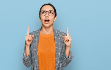 Young hispanic girl wearing business jacket and glasses amazed and surprised looking up and pointing with fingers and raised arms. 