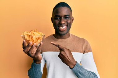 Young african american man holding bowl with potato chip smiling happy pointing with hand and finger 