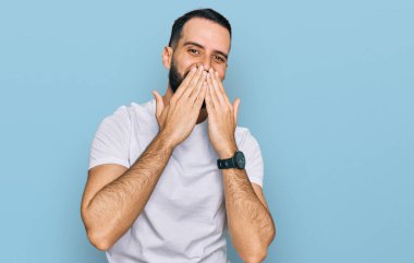 Young man with beard wearing casual white t shirt laughing and embarrassed giggle covering mouth with hands, gossip and scandal concept 