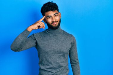 Arab man with beard wearing turtleneck sweater smiling pointing to head with one finger, great idea or thought, good memory 