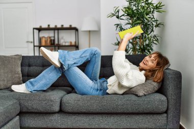 Evdeki kanepede uzanmış kitap okuyan beyaz bir kadın.