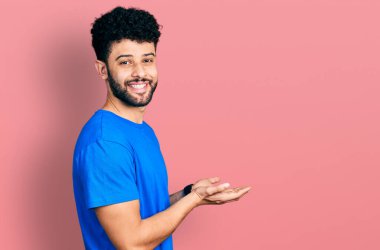Young arab man with beard wearing casual blue t shirt pointing aside with hands open palms showing copy space, presenting advertisement smiling excited happy 