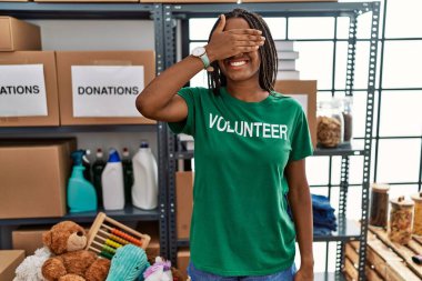 Young african american woman working wearing volunteer t shirt at donations stand smiling and laughing with hand on face covering eyes for surprise. blind concept. 