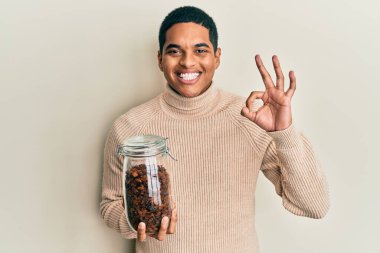 Genç, yakışıklı, elinde kuru üzümlerle dolu kavanoz tutan yakışıklı bir adam parmaklarıyla imza atıyor, gülümseyerek mükemmel bir sembol oluşturuyor. 