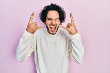 Handsome hispanic man wearing casual white sweater shouting with crazy expression doing rock symbol with hands up. music star. heavy music concept. 