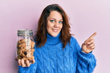 Young caucasian woman holding jar of chocolate chips cookies smiling happy pointing with hand and finger to the side 