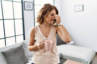 Orta yaşlı bir kadın evde kahve içiyor. Yorgun, burnu ve gözleri ovuşturup yorgun ve başı ağrıyor. Stres ve hüsran kavramı. 