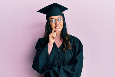 Young hispanic woman wearing graduation cap and ceremony robe looking confident at the camera with smile with crossed arms and hand raised on chin. thinking positive. 