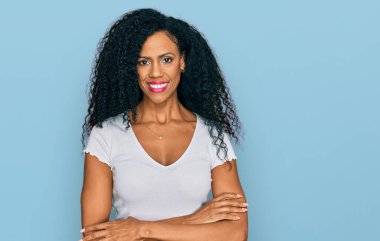 Orta yaşlı, beyaz tişört giyen, kollarını kavuşturup kameraya gülümseyen Afro-Amerikan kadın. pozitif kişi. 