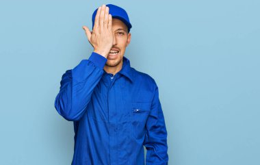 Bald man with beard wearing builder jumpsuit uniform covering one eye with hand, confident smile on face and surprise emotion. 