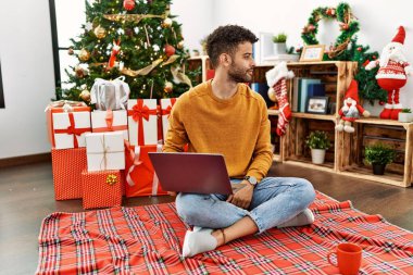 Arab young man using laptop sitting by christmas tree looking to side, relax profile pose with natural face and confident smile. 