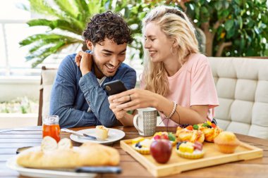 Genç çift kahvaltı yapıyor ve akıllı telefon kullanıyor terasta masada oturuyor..