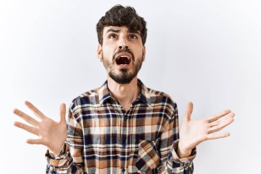 Hispanic man with beard standing over isolated background crazy and mad shouting and yelling with aggressive expression and arms raised. frustration concept. 