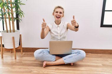 Young blonde woman using computer laptop sitting on the floor at the living room approving doing positive gesture with hand, thumbs up smiling and happy for success. winner gesture. 