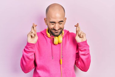 Young bald man wearing gym clothes and using headphones gesturing finger crossed smiling with hope and eyes closed. luck and superstitious concept. 