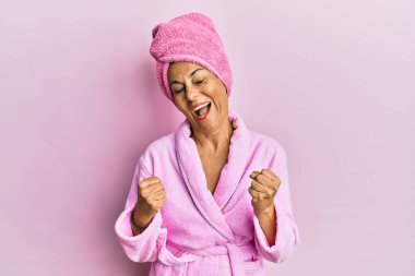 Middle age hispanic woman wearing shower towel cap and bathrobe very happy and excited doing winner gesture with arms raised, smiling and screaming for success. celebration concept. 