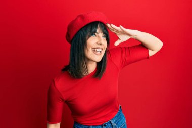 Young hispanic woman wearing french look with beret very happy and smiling looking far away with hand over head. searching concept. 