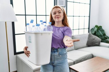 Elinde plastik şişelerle çöpleri geri dönüşüm sepetine koyan genç kızıl saçlı kadın neşeli gülümseyerek avuç içi yardım ve kabulü sunuyor.. 
