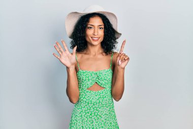 Young latin girl wearing summer hat showing and pointing up with fingers number six while smiling confident and happy. 