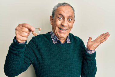 Handsome senior man with grey hair holding medical hearing aid celebrating victory with happy smile and winner expression with raised hands 