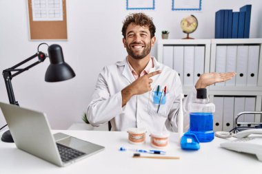 Young hispanic dentist man working at medical clinic amazed and smiling to the camera while presenting with hand and pointing with finger. 
