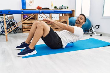 Young hispanic man patient smiling confident having back rehab session using fit ball at clinic