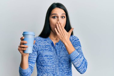 Young hispanic woman holding take away coffee covering mouth with hand, shocked and afraid for mistake. surprised expression 