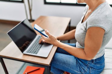 Evde akıllı telefon ve laptop kullanan orta yaşlı sarışın bir kadın.