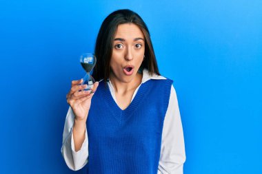 Young hispanic girl holding sand clock scared and amazed with open mouth for surprise, disbelief face 