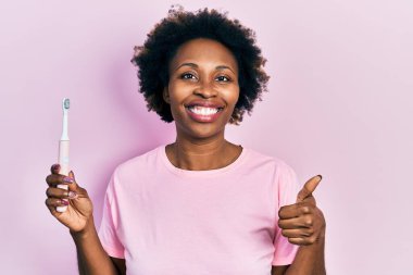Young african american woman holding electric toothbrush smiling happy and positive, thumb up doing excellent and approval sign 