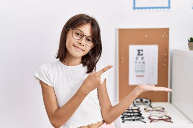 Young hispanic girl wearing glasses amazed and smiling to the camera while presenting with hand and pointing with finger. 