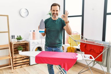 Middle age man with beard ironing clothes at home doing ok sign with fingers, smiling friendly gesturing excellent symbol 