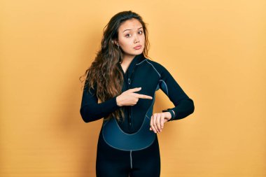 Young hispanic girl wearing diver neoprene uniform in hurry pointing to watch time, impatience, upset and angry for deadline delay 