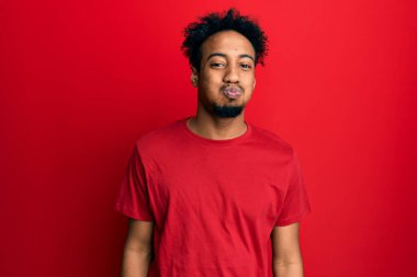 Young african american man with beard wearing casual red t shirt puffing cheeks with funny face. mouth inflated with air, crazy expression. 