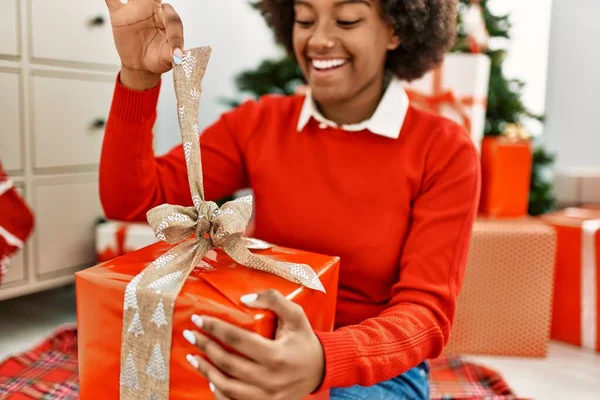 Genç Afro-Amerikan kadın boks hediyesini açıyor evde Noel ağacının yanında oturuyor.