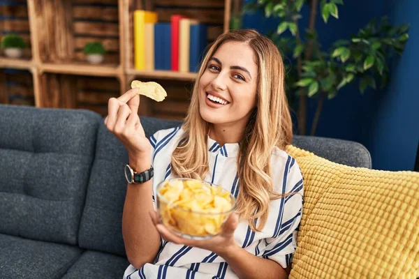 Young Blonde Woman Eating Chips Potatoes Sitting Sofa Home — Stockfoto