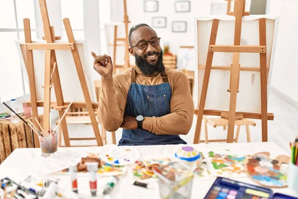 African american artist man at art studio with a big smile on face, pointing with hand and finger to the side looking at the camera. 