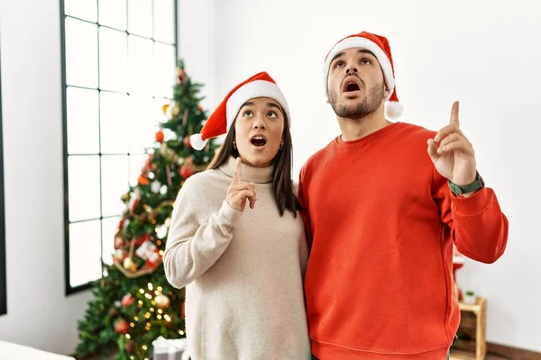 Young Hispanic Couple Standing Christmas Tree Amazed Surprised Looking Pointing — Stok fotoğraf