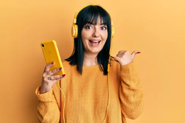 Young Hispanic Woman Using Smartphone Wearing Headphones Pointing Thumb Side — 스톡 사진