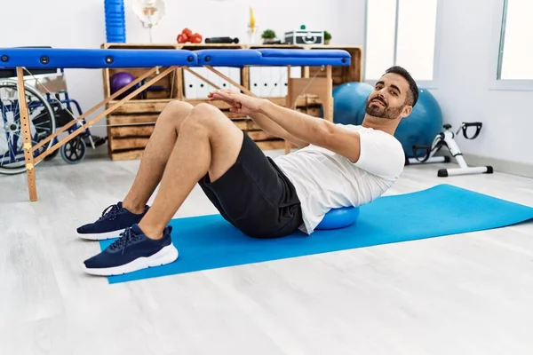 Young hispanic man patient smiling confident having back rehab session using fit ball at clinic