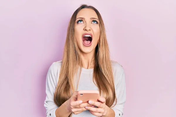 Young Blonde Girl Using Smartphone Typing Message Angry Mad Screaming — Fotografia de Stock