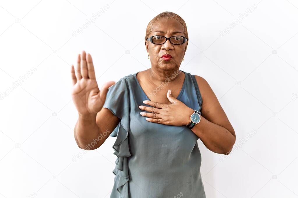 Mature hispanic woman wearing glasses standing over isolated background swearing with hand on chest and open palm, making a loyalty promise oath 
