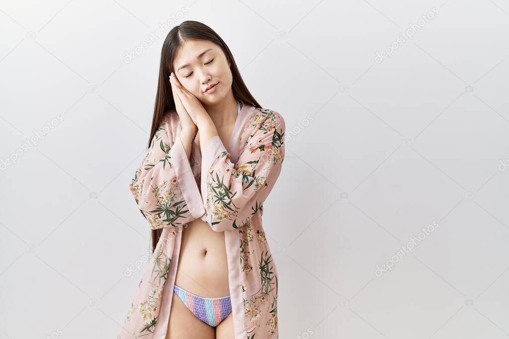 Young asian woman wearing bikini an floral kimono sleeping tired dreaming and posing with hands together while smiling with closed eyes. 