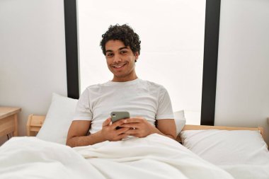 Genç İspanyol adam yatak odasındaki yatakta akıllı telefon kullanıyor..