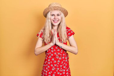 Beautiful caucasian woman with blond hair wearing summer hat praying with hands together asking for forgiveness smiling confident. 